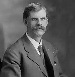A man with dark hair and a prominent mustache wearing a black jacket, patterned tie, and white shirt