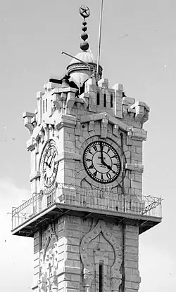 Black-and-white photograph of a stone clock tower with four sides. On the sunny side, a clock face with Roman numerals. On the shaded side, a clock face with Arabic numerals. The clock hands are in different positions on each face. There is a spire with spheres and a crescent and star on the top of the tower.