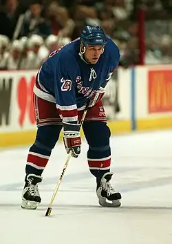 A Caucasian hockey player. He wears a dark blue jersey and helmet. He is bent over at the waist leaning on his stick.
