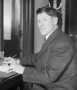 Black-and-white image of Jim Thorpe sitting down and writing. He is a Native American man with a Western haircut and wearing a dark suit.