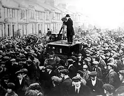 A person stand atop a platform operating a camera whilst surrounded by a large crowd on a street lined with buildings. The scene depicts John Maclean’s casket being removed from his home.