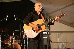 Bald Middle Eastern man wearing glasses and black shirt and pants, standing on stage, holding a guitar, and singing into a microphone in front of a drum kit.