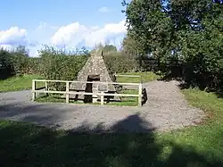A pyramidal stone structure stands in a small clearing surrounded by small trees and bushes. The structure, enclosed by a fence, has an opening in the front.