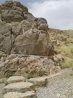 Photograph of an outdoor scene. In first term, a gravel floor and four stepping stones on it. At the end of the stones, a rock outcrop. There is a flat level about the height of the viewer. Upon it, the rock rises. The top of the outcrop is shaped as the reclining Hercules. Behind him, the rock rises more. Behind it, the blue sky with some clouds. To the right, the slope is covered with grass.