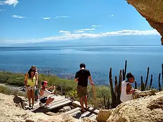 Five people stand and sit on a cliff, overlooking vegetation below and a large lake beyond. The lake stretches the full width of the photo, and the shoreline on the other side is barely visible.