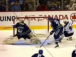 An ice hockey goaltender stretches across the net to get into position as a teammate stands to the right of the net, keeping an opposing forward away.