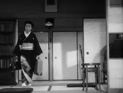 A middle-aged Japanese woman, wearing a kimono and carrying a suitcase in her left hand and a valise in her right, in the process of walking around the perimeter of a small room. There is a bookcase on the left and a straight-backed chair on the right and screens and a ceiling lamp in the near background.