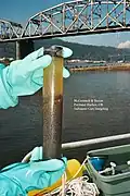 A diver's blue-gloved hands holds a cylindrical sediment core. The core is inside a plastic tube through which dark sediments and murky water can be seen. Image text reads "McCormick & Baxter Portland Harbor, OR Sediment Core Sampling". In the background there is the deck of a boat, water, a bridge, and a forested ridge.