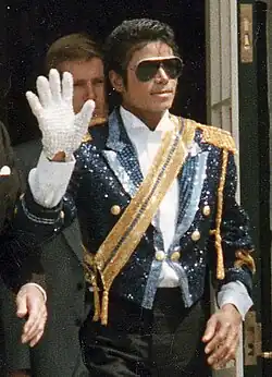 Michael Jackson in a blue suit with a gold diagonal strike, and a white shirt underneath. He has white gloves, waving with his right hand. He is wearing sunglasses.