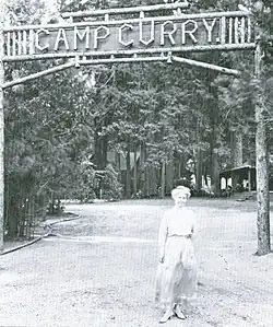 Woman in a long dress in front of a sign across a road. Wooden letters read "Camp Curry".
