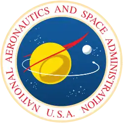 A blue sphere with stars, a yellow planet with a white moon; a red chevron representing wings, and an orbiting spacecraft; surrounded by a white border with "NATIONAL AERONAUTICS AND SPACE ADMINISTRATION U.S.A." in red letters