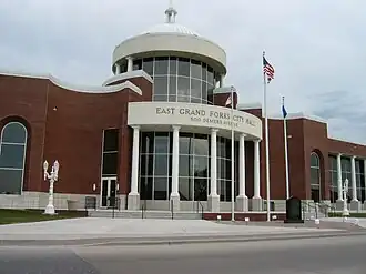 A red brick building with a glass lobby. A memorial sign and flags are located in front of the building.