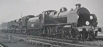A side-and-front view of a 2-6-0 steam locomotive coupled to a breakdown crane in a yard. The locomotive features smoke deflector plates either side of the boiler and there are white route discs on the front.