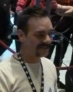 A man with dark hair and a handlebar mustache looking and smiling to the right of the camera.