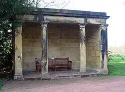 A open stone structure with four columns supporting a lintel. Inside the loggia is a wooden seat.