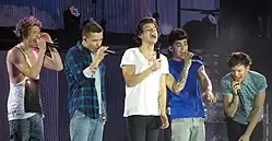 Five teenage boys standing next to each other and singing on stage. They all wear jeans, and everyone except the boy standing on the far left wears a t-shirt. The boy on the far-left wears a tank top. From left to right: Niall Horan, Liam Payne, Harry Styles, Zayn Malik, and Louis Tomlinson.