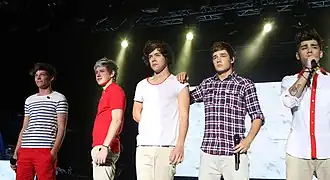 Five teenage boys stand next to each other on a stage. From left to right: Louis Tomlinson, Niall Horan, Harry Styles, Liam Payne, and Zayn Malik. Tomlinson wears bright red trousers and a white t-shirt with horizontal black stripes, and smiles while gazing at the audience. Horan, Styles, Payne, and Malik all wear identical cream trousers. Horan wears a red polo shirt. Styles wears a white t-shirt with a red rim around its neckline. Payne wears a checkered dress shirt. Malik wears a white dress shirt, with red lining around the buttons and collar. Payne has his right hand on Styles' shoulder, and Malik is the only member visibly singing.