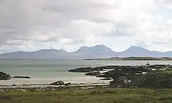 A small sandy beach lies beyond a grassy foreshore. In the distance, the outline of a range of brown and grey hills is visible under cloudy skies.