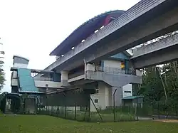 An elevated station (Teck Lee) with a distinctive curved roof. The station is surrounded by a fence as it stood among grassy areas and some trees, with the elevated train tracks extending on either side.