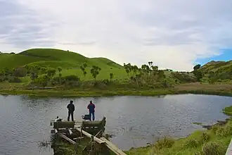 Two people on a dock in a small body of water, surrounded by rolling green hills and shrub