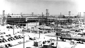 Expansive view of a construction site with lots of parked cars, scaffolding and cranes. There are a number of demountables in the foreground.