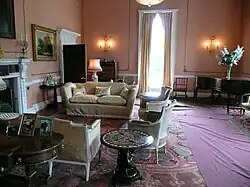 Drawing room with high ceilings and tall window having biscuit coloured walls with white raised mouldings, soft furnishings and piano in corner