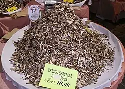 A heaped pile of dried sliced mushrooms on a large plate. A sign near the back of the plate reads "Fungo di Borgotaro I.G.P."; another sign at the front reads "Prezzo speciale di Fiera. 3 Etti 18,00". An electronic scale is partially visible on a table behind the mushrooms