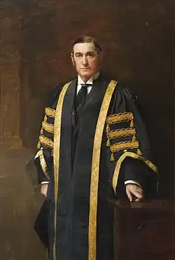 Three quarter length standing portrait, the subject looking directly at the viewer, while turned slightly to left. One hand rests on a low wooden table and he wears gowns which are black with a gold trim and details. Standing against a brown background.