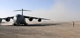 Colour photo of a grey military aircraft on a dusty airstrip. A person is standing to the right of the aircraft.
