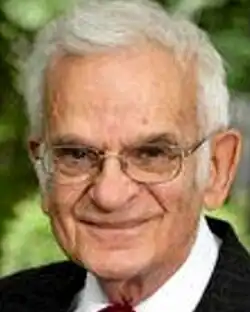 Colour press shot of elderly, smiling, clean-shaven and bespectacled man, with white hair swept back.