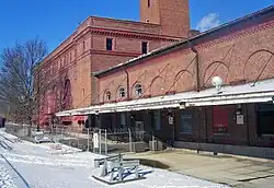 A brick building with snow-covered ground and a bare tree at the lower left. The portion of the building nearer the camera, on the right, has large rounded indentations in it and a metal roof hung from its side by cables over a bare concrete surface. The portion on the left, farther away, has been fenced off. It is taller, with a large square section rising to the top of the image in the center.