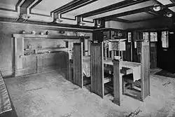 Interior of the dining room as seen in 1911. The dining room chairs have high, flat backs. The chairs surround a table with lamps at its corners.