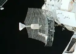 A rectangular dish shape of scaffolding covered in transparent sheeting, with a white insulation-covered radio receiver and support projecting from the centre. The blackness of space serves as the backdrop.