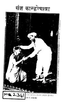 Cover-page of a script titled Saint Kanhopatra (संत कान्होपात्रा) in Marathi, with a photograph showing a woman dressed in a sari kneeling with palms pressed together before a man. The man, dressed in a white dhoti-kurta, is bent forward and seems to be blessing her with his wight hand placed over her head.