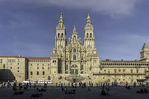 A very large cathedral in Santiago de Compostela from the front, occupying the whole image with some people sitting and standing in front on cobblestones.