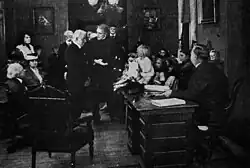 Black and white film still showing a dramatic scene in a crowded room
