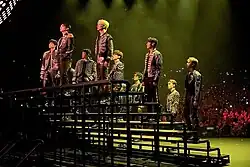 The members of Seventeen standing on a metal structure on a stage at the beginning of a concert