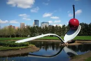 A large gray spoon straddles a shallow pond. On the tip of the spoon is a stylized red cherry with a black stem. The sculpture is surrounded by lawn and in the background, coniferous trees, behind which several skyscrapers stand. The sky is blue with several clouds.