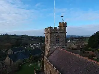 The image shows St John the Baptist church, Broadwindsor, from an aerial perspective taken using a drone. The church tower features prominently in the middle of the image, with views of the countryside and village of Broadwindsor in the background