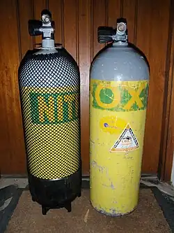 Two cylinders stand next to each other. On the left is a round-bottomed 15-litre steel cylinder with a plastic boot, and on the right a flat-bottomed 12.2-litre aluminium cylinder without boot. Both cylinders are the same outside diameter (203 mm), but the smaller-volume aluminium cylinder is slightly higher than the larger-volume steel cylinder, even though the steel cylinder is standing on a boot and has a rounded bottom.