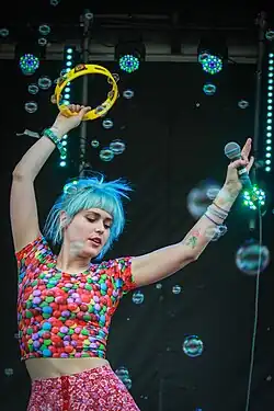 Emily Nokes, a blue-haired white woman, waves a tambourine while dancing in a cloud of bubbles
