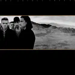 A landscape monochrome photograph of U2 in the desert sits in the center of a black background. U2 are standing on the left half of the photograph, with a mountain range on the right half. Tiny gold text reading "THE JOSHUA TREE U2" is stretched across the top of the black background.