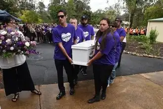 A group of six people dressed in purple carry a white coffin from concrete onto a footpath, next to a woman in a white shirt holding a bouquet.