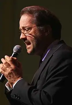 A middle-aged man wearing glasses and a dark suit, talks to a microphone.