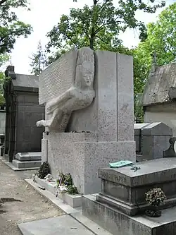A stone sculpture of a winged sphinx-like figure on a tomb, carved in an angular, modernist style