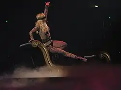 Image of a blond woman. Her right hip is resting on a black divan, with her right leg extended to the end of the divan. Her left hand is extended in the air, while her right hand is grabbing the top of the divan. She is wearing a flesh-colored body suit with tattoos and nipple tassles. She is sporting a black blindfold and singing into a wireless microphone. The divan is surrounded by smoke.