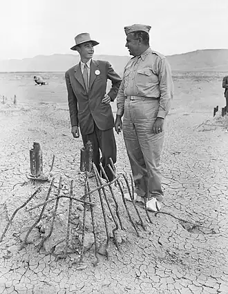 A man smiling in a suit in suit and one in a uniform chat around a pile of twisted metal.
