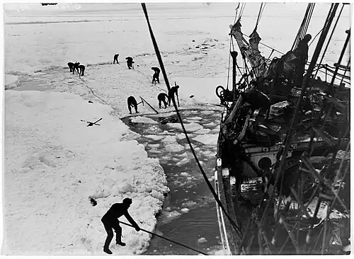 Men with digging tools removing ice surrounding the ship's hull, creating an icy pool of water