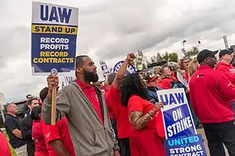 An outdoor strike rally. UAW members of all races are raising their fists, wearing bright red T-shirts, and holding picket signs with following messages: "UAW On Strike", "Stand Up / Record Profits / Record Contracts".
