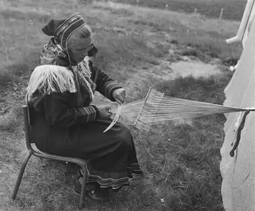 A Sámi weaver doing inkle weaving on a backstrap loom with a rigid heddle. She seems to be using a hollow half-bone as a beater and as a race for a bobbin. Norway, 1956.